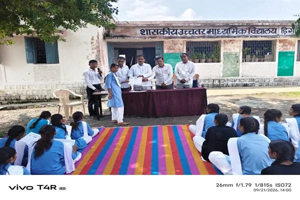 स्कूल परिसर में मेज के पीछे खड़े अतिथि एक छात्रा को प्रमाण-पत्र देते हुए, सामने बैठीं छात्राएं