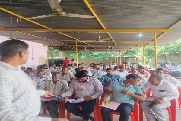छायादार परिसर में लाल कुर्सियों पर बैठकर कागजों में जानकारी दर्ज करते दुग्ध उत्पादक सदस्य