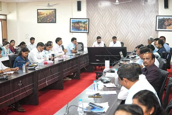 सभाकक्ष में लंबी मेजों के दोनों ओर बैठे अधिकारी बैठक में दस्तावेज और स्क्रीन देखते हुए