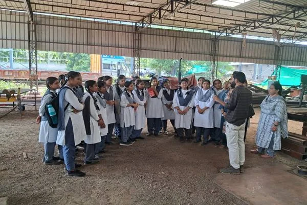एक औद्योगिक शेड के भीतर सफेद-नीली स्कूल यूनिफॉर्म पहने छात्राओं का समूह खड़ा है, सामने खड़े एक व्यक्ति उन्हें कुछ समझा रहे हैं और पास में एक महिला खड़ी हैं।