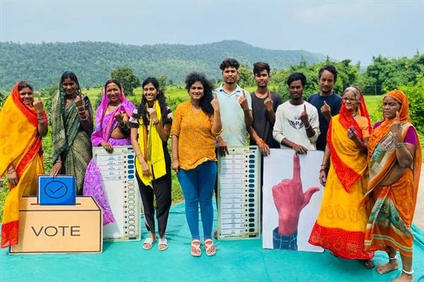 खेतों और पहाड़ियों के सामने खड़े ग्रामीण महिलाएं और युवा स्याही लगी उंगली दिखाते हुए, साथ में ईवीएम के मॉडल, 'VOTE' लिखा मतपेटी जैसा डिब्बा और स्याही लगी उंगली का बड़ा पोस्टर।
